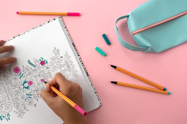 Flatlay with antistress coloring with flowers, pens, pencil case, cup of tea and female hands.