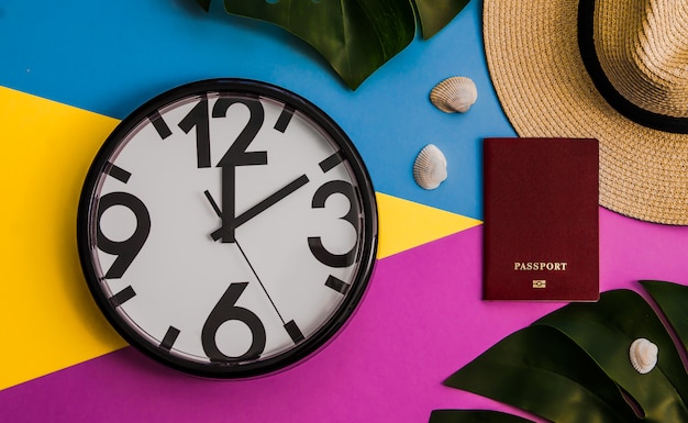 Flatlay with monstera leaves, clock, passport, straw hat on three tone background.