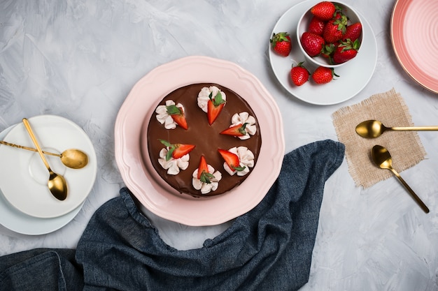Flatlay with vegan chocolate cake, strawberries and empty plates with gold spoons on cement background with copyspace
