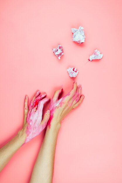 Flatlay with woman's hands covered in white and red paint and cramped paper on pink, creativity concept