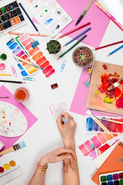 Flatlay with woman's hands holding brush, paletts, pencils, watercolors, colored paper and other stationary supplies, artist or designer workplace