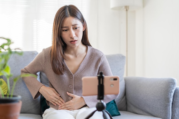 사진 헛배 부른 아시아 젊은 여성, 소녀 또는 환자의 손은 식중독, 복통 및 결장 문제, 위염 또는 설사로 고통받습니다. 스마트폰 개념에 대한 원격 의료 또는 원격 의료.