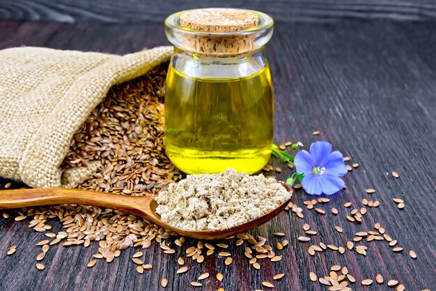 Foto farina di semi di lino in un cucchiaio, olio in un barattolo di vetro, fiore di lino blu, semi marroni in un sacchetto su uno sfondo di tavola di legno scuro