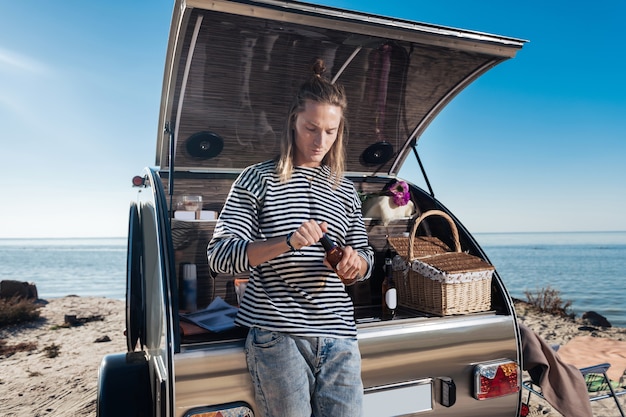 Flesje bier. Blondharige hipster die een gestreept overhemd draagt dat flesje bier houdt dat zich dichtbij zijn kleine compacte aanhangwagen bevindt