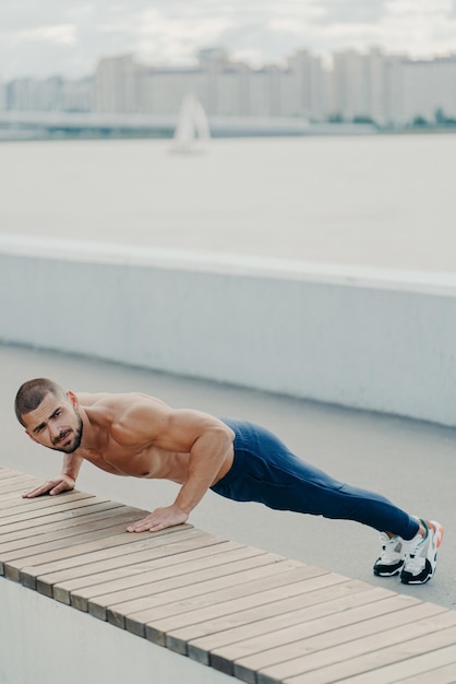 Flexibele fit man doet plank oefening op promenade heeft naakte torso geconcentreerd weg bereidt zich voor op workout praktijken yoga gekleed in sportkleding poseert buiten treinen in de ochtend Sportieve levensstijl