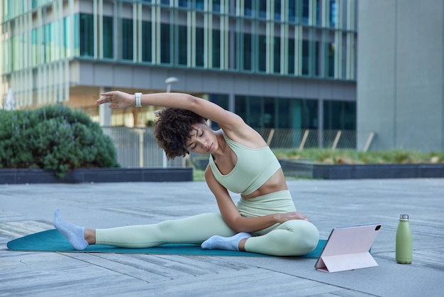 Flexibele slanke vrouw in trainingspak oefent yogahoudingen uit bij karemat kijkt naar instructievideo's op tablet leunt weg houdt arm omhoog