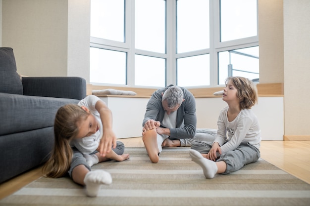 Flexibiliteit oefening. Betrokken jonge kinderen en volwassen grijsharige vader in comfortabele kleding doen flexibiliteit warming-up zittend op de vloer thuis