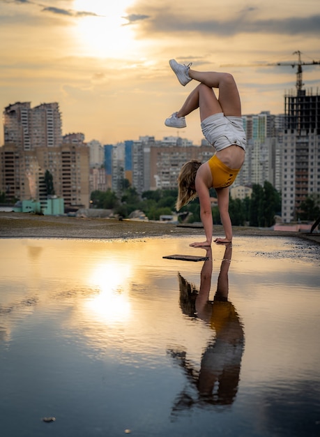 Flexible female gymnast doing handstand and calisthenic with reflection in the water on cityscape ba...