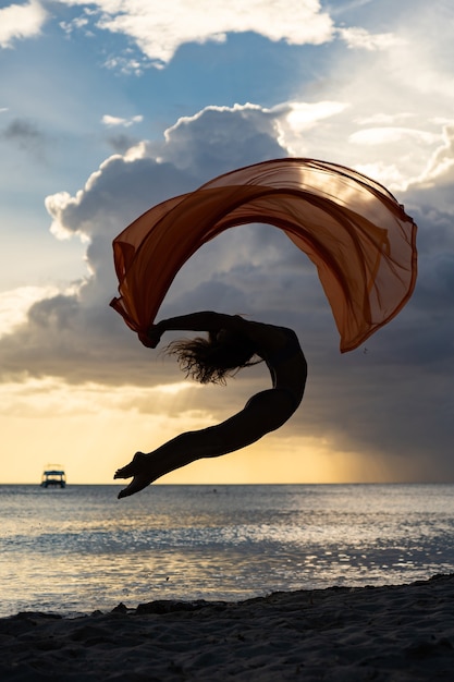 Donna in forma flessibile che salta con la seta durante il tramonto spettacolare con nuvole tempestose. concetto di individualità, creatività e fiducia in se stessi.