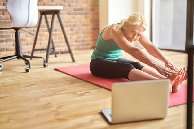 Flexible mature blonde woman in sportswear sitting on the floor stretching her body while watching