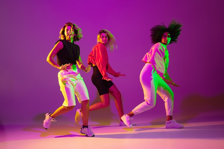 Photo フレキシブル。緑のネオンの光のダンスホールで紫ピンクの背景にスタイリッシュな服でヒップホップを踊るスポーティーな女の子。若者文化、動き、スタイルとファッション、行動。ファッショナブルな肖像画。