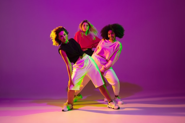 Flexible. Sportive girls dancing hip-hop in stylish clothes on purple-pink background at dance hall in green neon light. Youth culture, movement, style and fashion, action. Fashionable portrait.