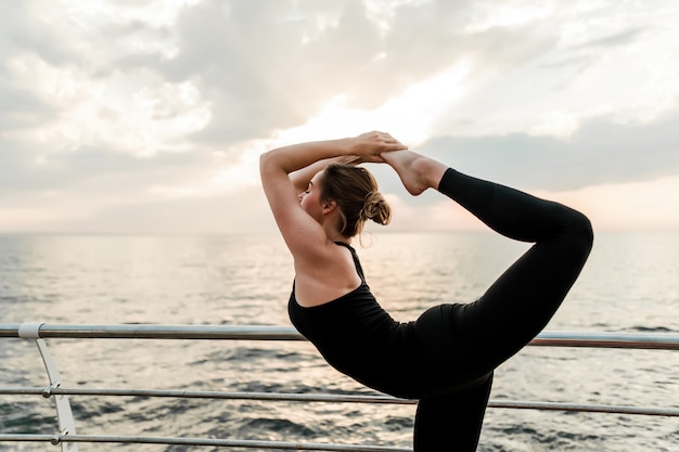 Flexible woman doing yoga asana near the sea on sunrise in the morning, practicing sport and fitness exercises