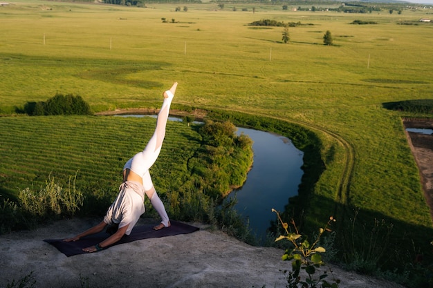 Flexible woman with stretching body performing ThreeLegged DownwardFacing Dog pose in evening outdoorson top of rock background of flowing river and green meadow on summer sunny day