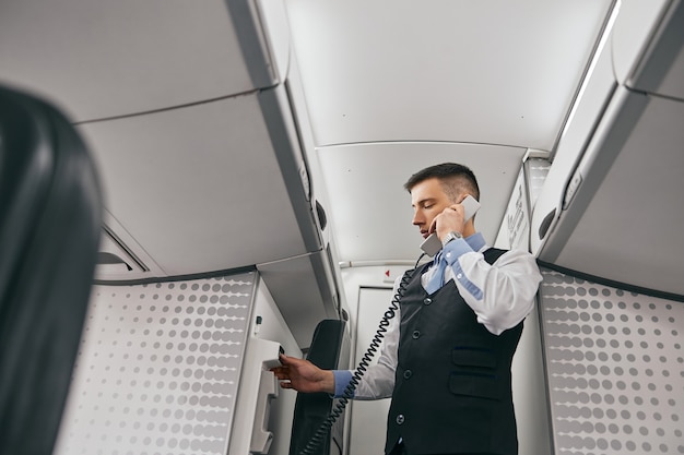 Flight attendant talk on board phone in passenger cabin of airplane jet. Modern plane interior. Bottom view of mixed race man wear uniform. Civil commercial aviation. Air journey concept