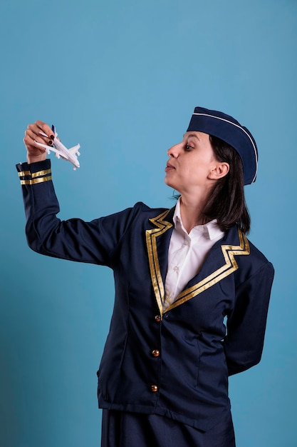 Flight attendant in uniform holding airplane model, aviation academy stewardess playing with plane. Air hostess looking at commercial jet toy, side view studio medium shot