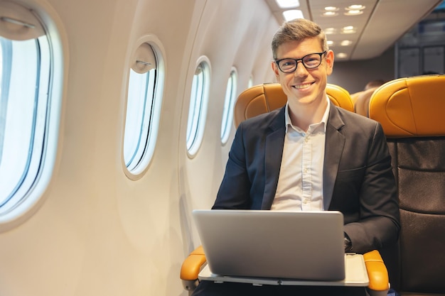 飛行中、フォーマルな服装と眼鏡をかけたビジネスマンが、インターネットアクセスと出張のコンセプトを備えたラップトップコンピューターサービスで作業している間、飛行機の窓の外を眺めます。