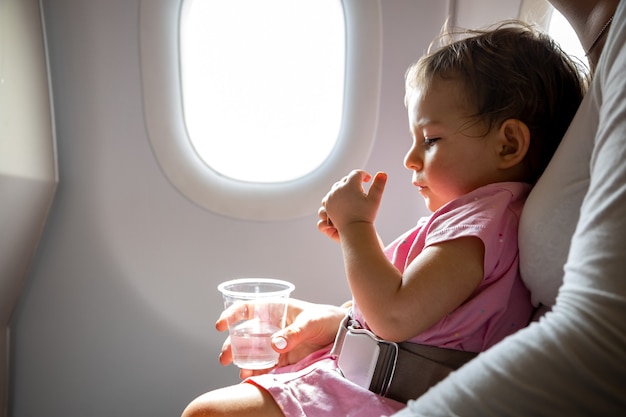 flight with infant. toddler sits on mom’s lap fastened with a special belt in airplane before porthole.