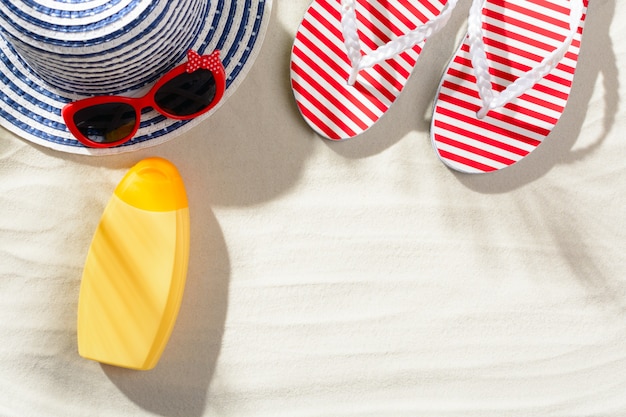 Flip flops, sunscreen, sunglasses and hat on the sand, top view