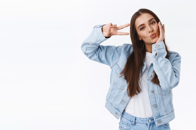 Flirterig en schattig, kokette brutale vrouw in trendy outfit, ontroerend wang kantelend hoofd vrouwelijk, vrede of overwinningsteken in de buurt van oog tonend als positieve vibes naar publiek, witte achtergrond