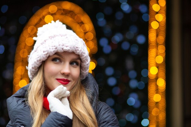 Flirterige blonde vrouw met rode lippenstift met een gebreide muts die op straat poseert op een achtergrond met slingers met bokeh