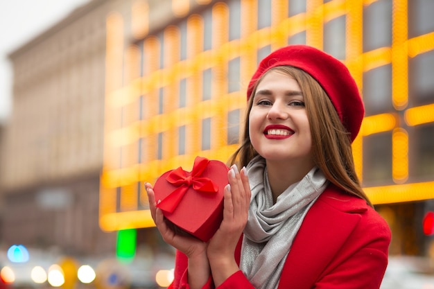 Flirterige schattige jonge vrouw met een hartvormige Valentijnsdag geschenkdoos met een strik op een wazige lichten achtergrond. Lege ruimte