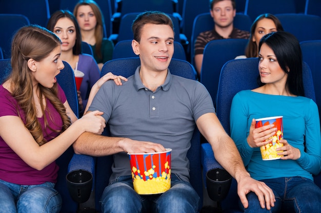 Flirting at the cinema. Young man sitting together with his girlfriend and holding hand on another woman knee while watching movie at the cinema