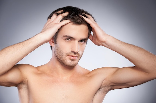 Flirty handsome. Portrait of handsome young beard man looking at camera and touching his hair with hands while standing isolated on grey background