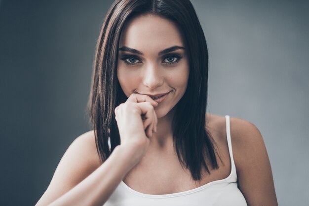 Flirty seductress. Attractive young woman holding hand on chin and looking flirty while sitting against grey wall