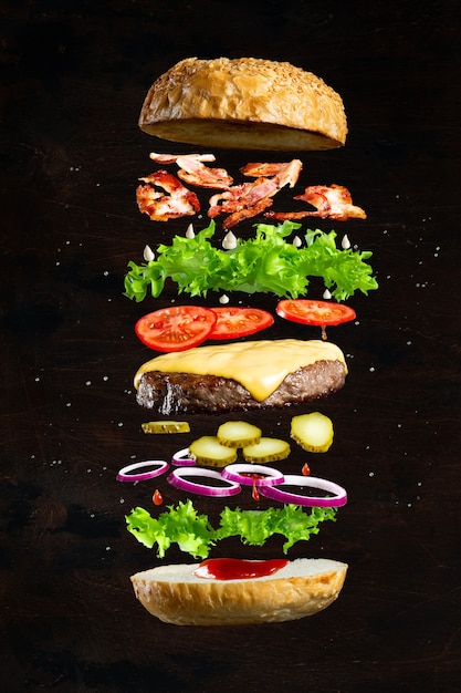 Floating burger isolated on black wooden background.\
ingredients of a delicious burger with ground beef patty, lettuce,\
bacon, onions, tomatoes and cucumbers