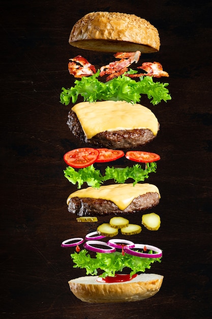 Floating burger isolated on black wooden background in men hands. Ingredients of a delicious burger with ground beef patty, lettuce, bacon, onions, tomatoes and cucumbers