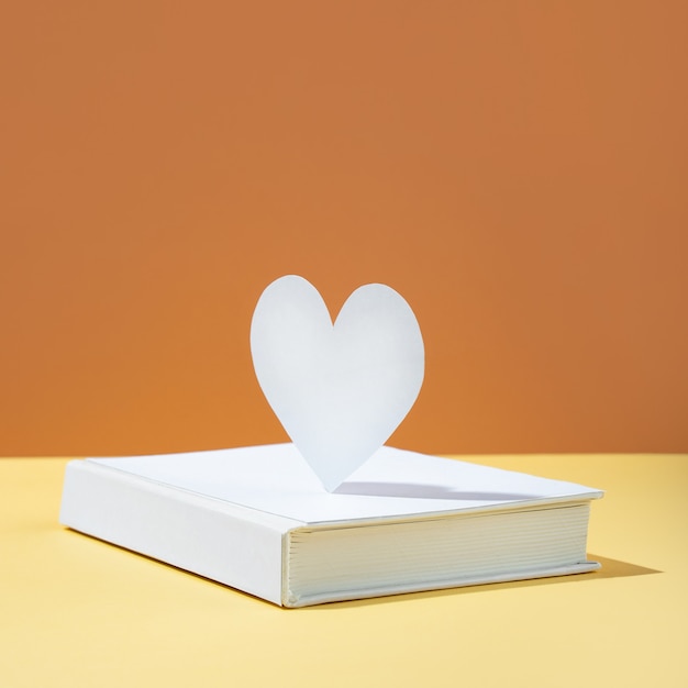 Floating cute paper heart and white cover book on bold color\
yellow and orange-brown background. modern photo. concept national\
book lovers day. happy holydays. romantic, love storytelling. copy\
space