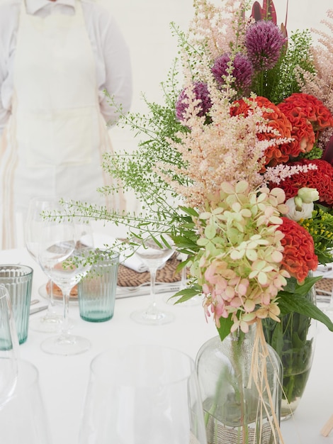 Floral arrangement on wedding table Glasses and cups on white tablecloth and unrecognizable waitress in white apron in the background out of focus