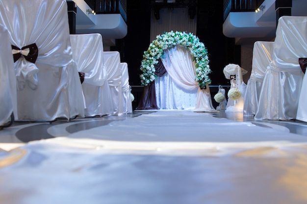 Photo floral bridal bower above a small altar with decorative white chairs tied in linen and bows viewed low angle along the aisle at a wedding venue