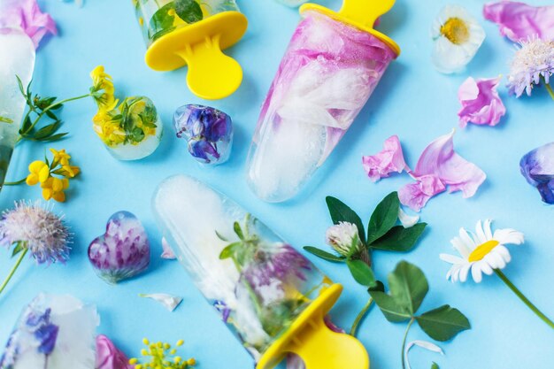 Цветочные леденцы Замороженное фруктовое мороженое и кубики льда из разноцветных полевых цветов на синем фоне плоско лежали со свежими летними цветами Привет летняя концепция Освежающие веганские сладости