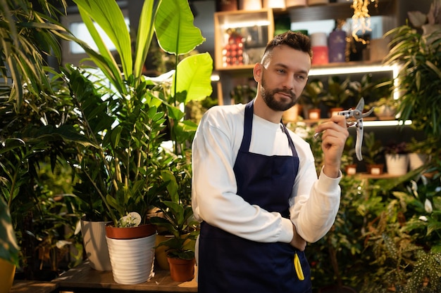 Florist gardener in a shop selling potted plants stands among flowers and holds a pruner in his