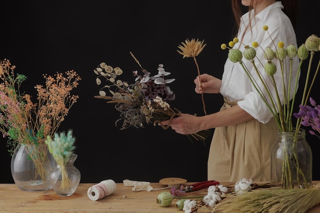 Florist makes a bouquet of dried flowers at a wooden table on a plain dark background with copyspace