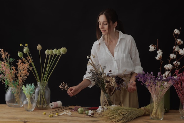 Florist makes a bouquet of dried flowers at a wooden table on a plain dark background with copyspace