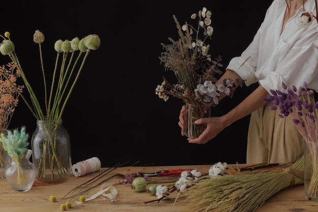 Florist makes a bouquet of dried flowers at a wooden table on a plain dark background with copyspace