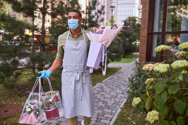 Флорист в маске гуляет на свежем воздухе с полиэтиленовыми пакетами с цветочными горшками и бумажным пакетом с букетом