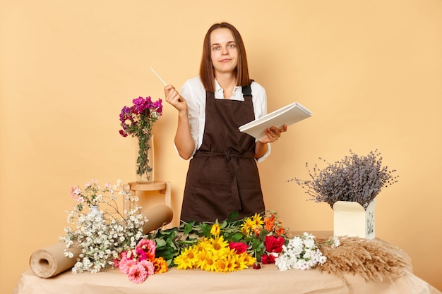 Foto spazio di lavoro creativo del fiorista belle composizioni di fiori fiori freschi in vendita donna fiorista incerta con scheda che scrive e fa appunti ordine al fiorista contro parete beige