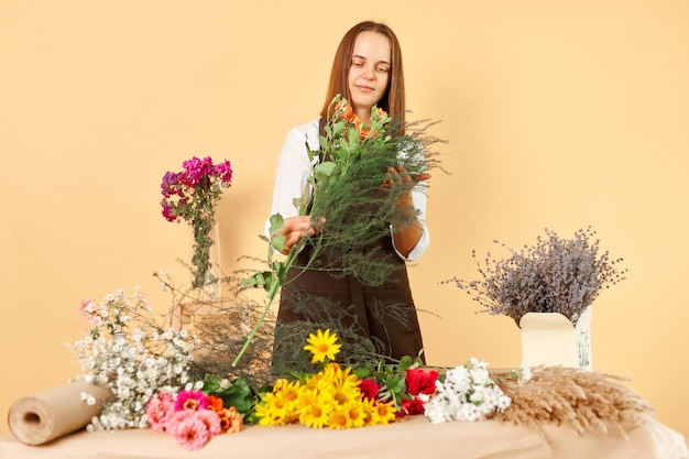 Florist's job Bunch of flowers Botanical delivery Gardener's occupation Floral arrangement Positive dark haired woman wearing brown apron working with beauty posing isolated over beige background