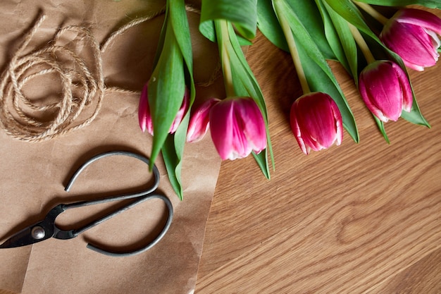 Posto di lavoro floristico con carta artigianale, spago disposizione di bouquet di tulipani rosa su tavola di legno, hobby, fai da te, concetto di regalo primaverile, dall'alto.