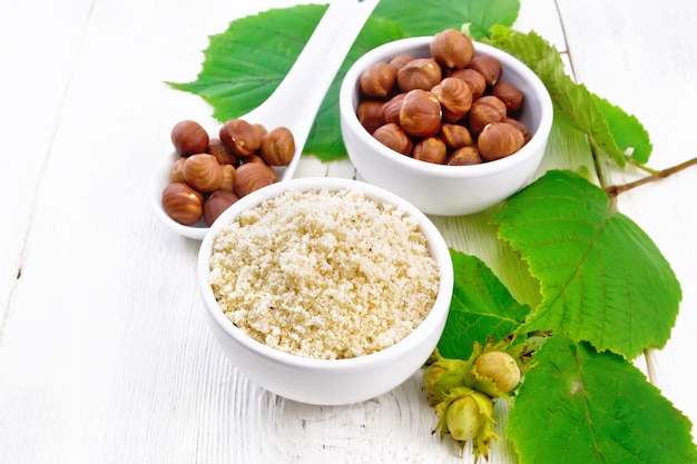 Flour and hazelnuts in two bowls, a spoon with peeled walnut kernels and a branch of filbert with leaves on the background of light wooden board