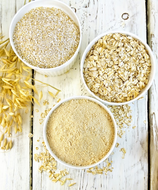 Flour oaten, bran and oat flakes in three white bowls, stalks on the background of the wooden planks from above
