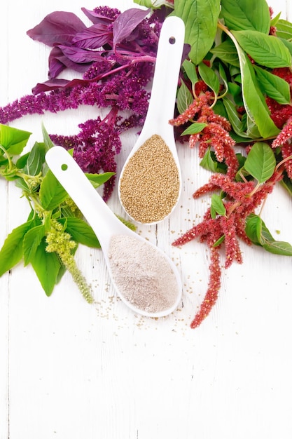 Flour and seeds amaranth in two spoons, multicolored flowers and leaves of a plant on the background of light wooden board from above