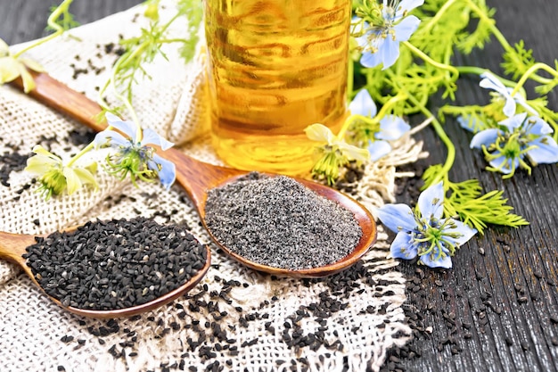 Flour and seeds of Nigella sativa in two spoons on a burlap oil in the bottle and twigs of kalingini with blue flowers and green leaves on wooden board background