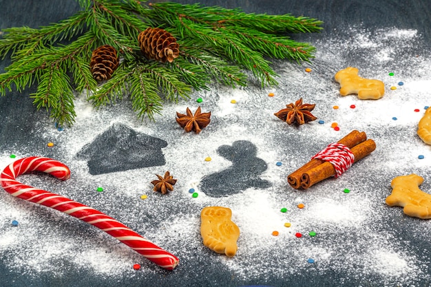 Flour silhouette of cookies on dark background among Christmas tree branches, cones, star anise, cinnamon and candy cane. 