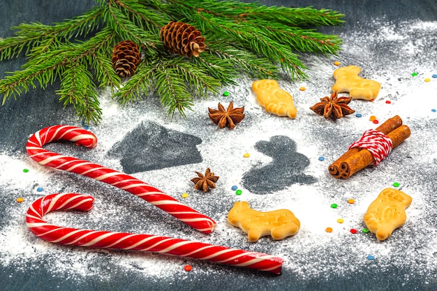 Flour silhouette of cookies on dark background among Christmas tree branches, cones, star anise, cinnamon and candy cane. 
