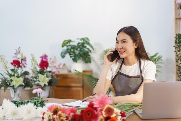 Concetto di negozio di fiori fiorista femminile che parla con il cliente sullo smartphone e prende nota sul notebook